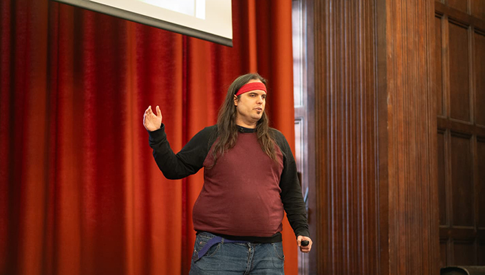 Photo of Mike Scrase, a person with long hair wearing a red headband and a Vans jumper, gesturing to a projector screen on stage at Bodies 2.