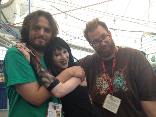 A photo of Mike Scrase, a non-binary person with way, medium long brown hair and a beard. Next to him is Ray Friesen, a man with short light brown hair, glasses and a beard. With them is a woman with black hair and lipstick, dressed in goth clothes.