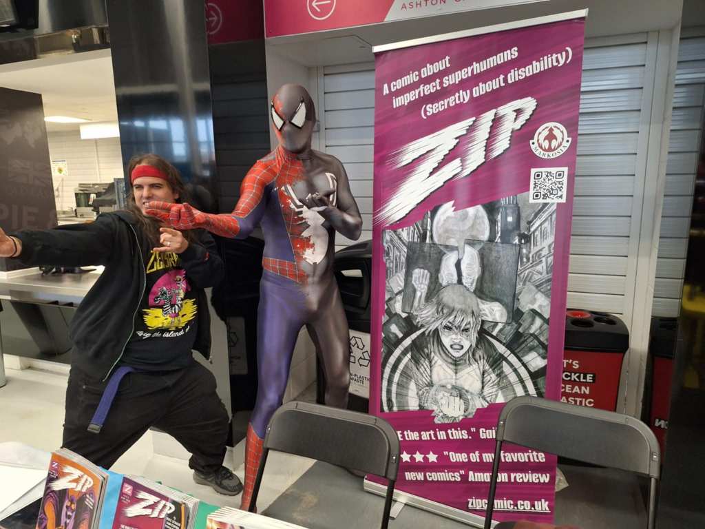Photo of writer Mike Scrase doing the 'thwip fingers': the gesture Spider-Man makes when he shoots a web (of any size), next to a Spider-Man cosplayer behind our convention table.